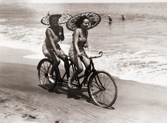ca. 1910, USA --- Two women in swimwear enjoy a tandem bike ride down the beach with parasols in hand. --- Image by © Underwood & Underwood/Underwood & Underwood/Corbis